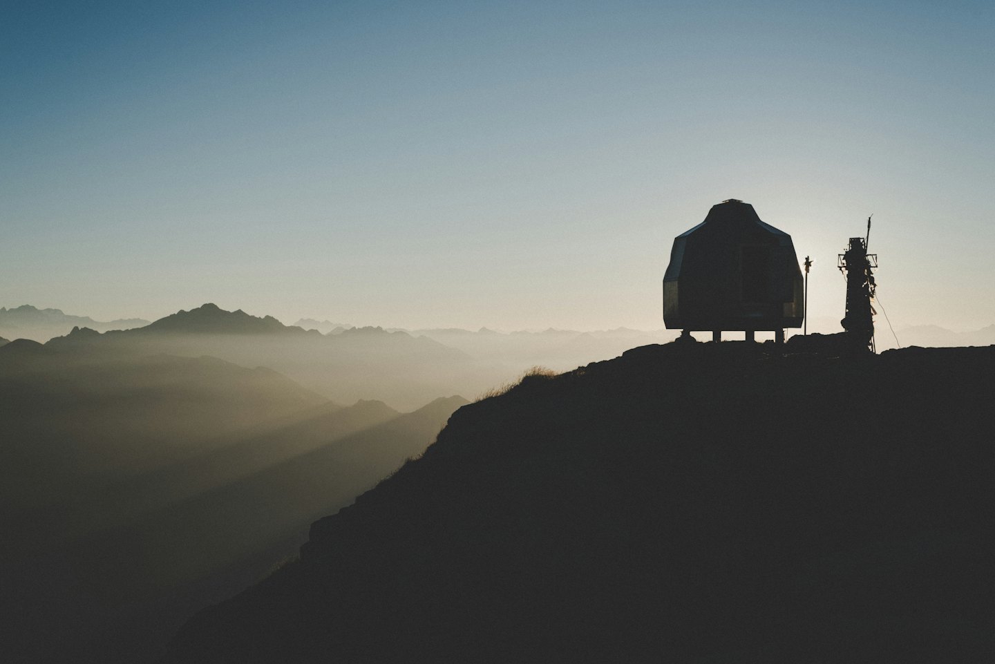 Hut and antenna silhouetted against hazy mountains at sunrise.